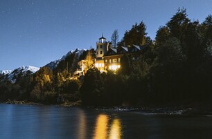 Un edificio grande e iluminado con una torre, rodeado de árboles y montañas nevadas, que se refleja en el agua.