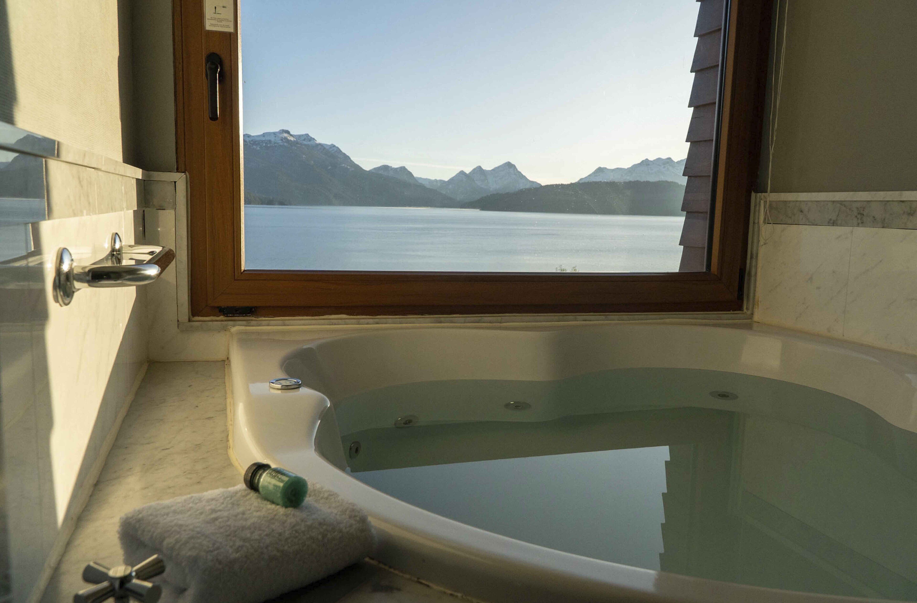 Uma casa de banho com banheira, janela e vista para um lago e montanhas.