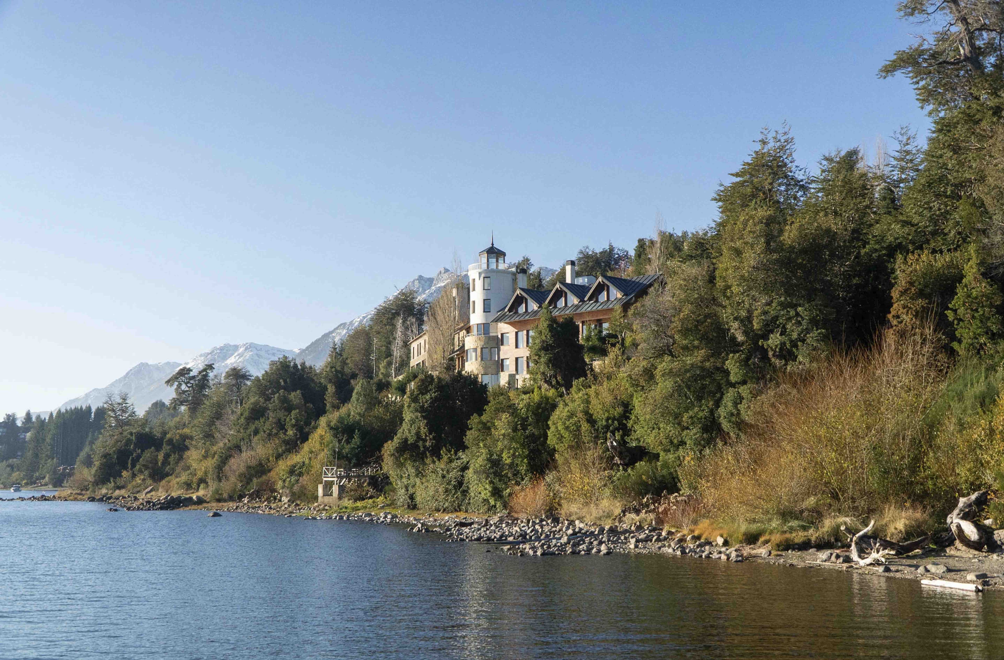 Una propietat a la vora d'un llac, amb un edifici gran de diverses plantes envoltat per un bosc dens i muntanyes.