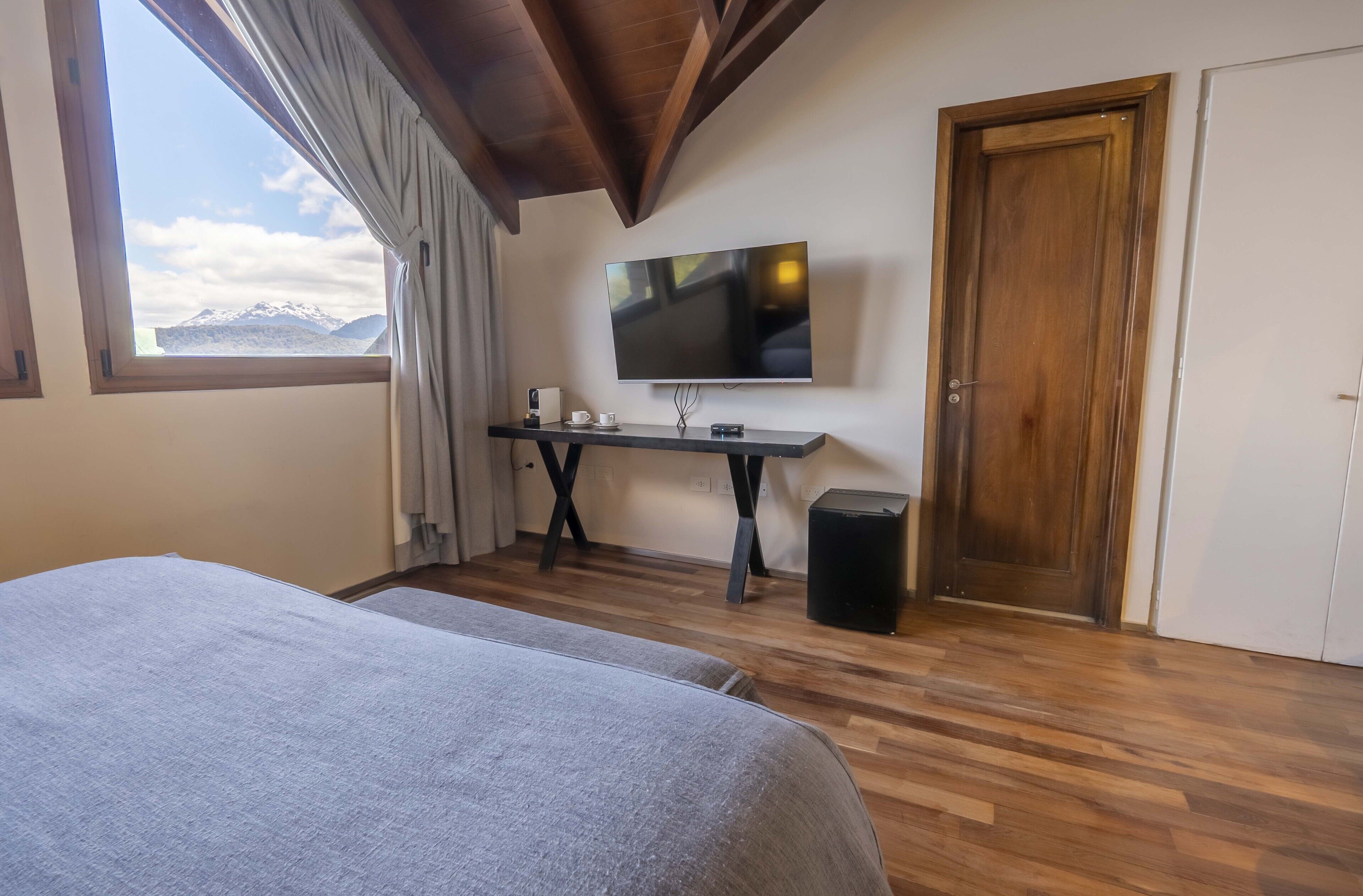 A hotel room with a bed, a desk with a TV, a window with a mountain view, and a door.
