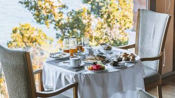 A breakfast table set with various dishes, including croissants, fruit, and beverages, overlooking a scenic view of trees and a body of water.