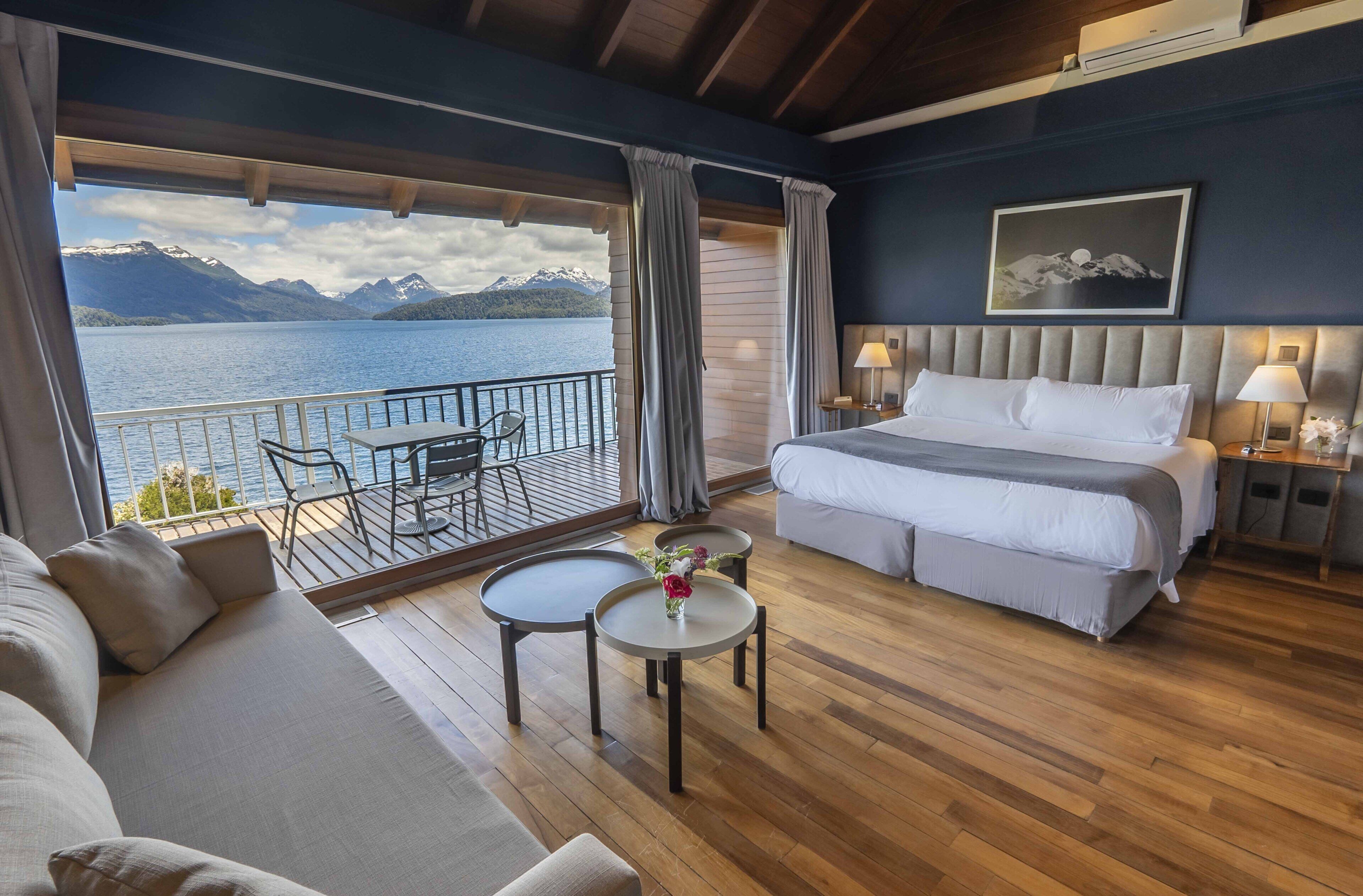 Una habitación con cama, sofá, una mesita con un florero, y un balcón con vistas a un lago y montañas.