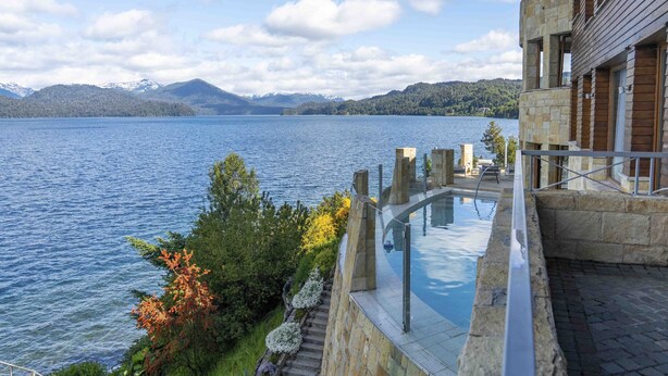 A modern building with a glass infinity pool overlooking a lake, stone architecture, and a mountainous backdrop. - El Faro Boutique Hotel & Spa by DON (Villa La Angostura)
