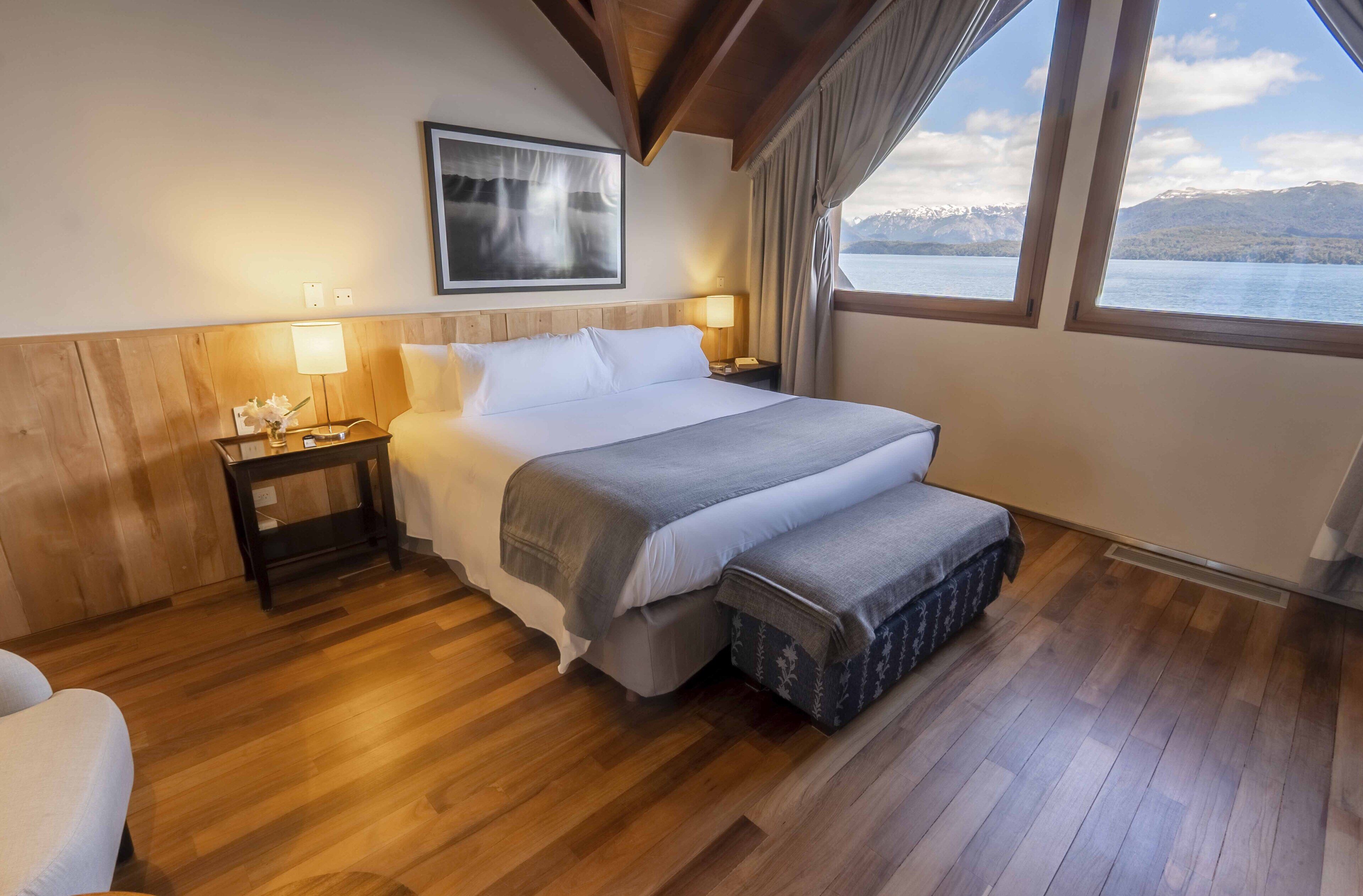 A bedroom with a wooden floor, a bed with a gray blanket, a nightstand with a lamp, and a window with a view of mountains and a lake.