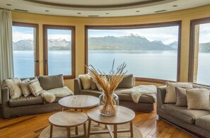 A living room with a view of a lake and mountains. There are two sofas and two round tables. - El Faro Boutique Hotel & Spa by DON (Villa La Angostura)