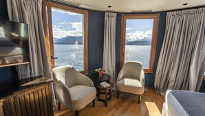 A room with a bed, two armchairs, a small table with a vase, and a TV on a cabinet. Large windows offer a view of a lake and mountains. - El Faro Boutique Hotel & Spa by DON (Villa La Angostura)