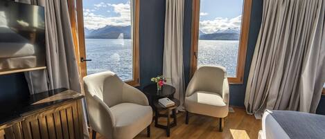 A room with a bed, two armchairs, a small table with a vase, and a TV on a cabinet. Large windows offer a view of a lake and mountains.