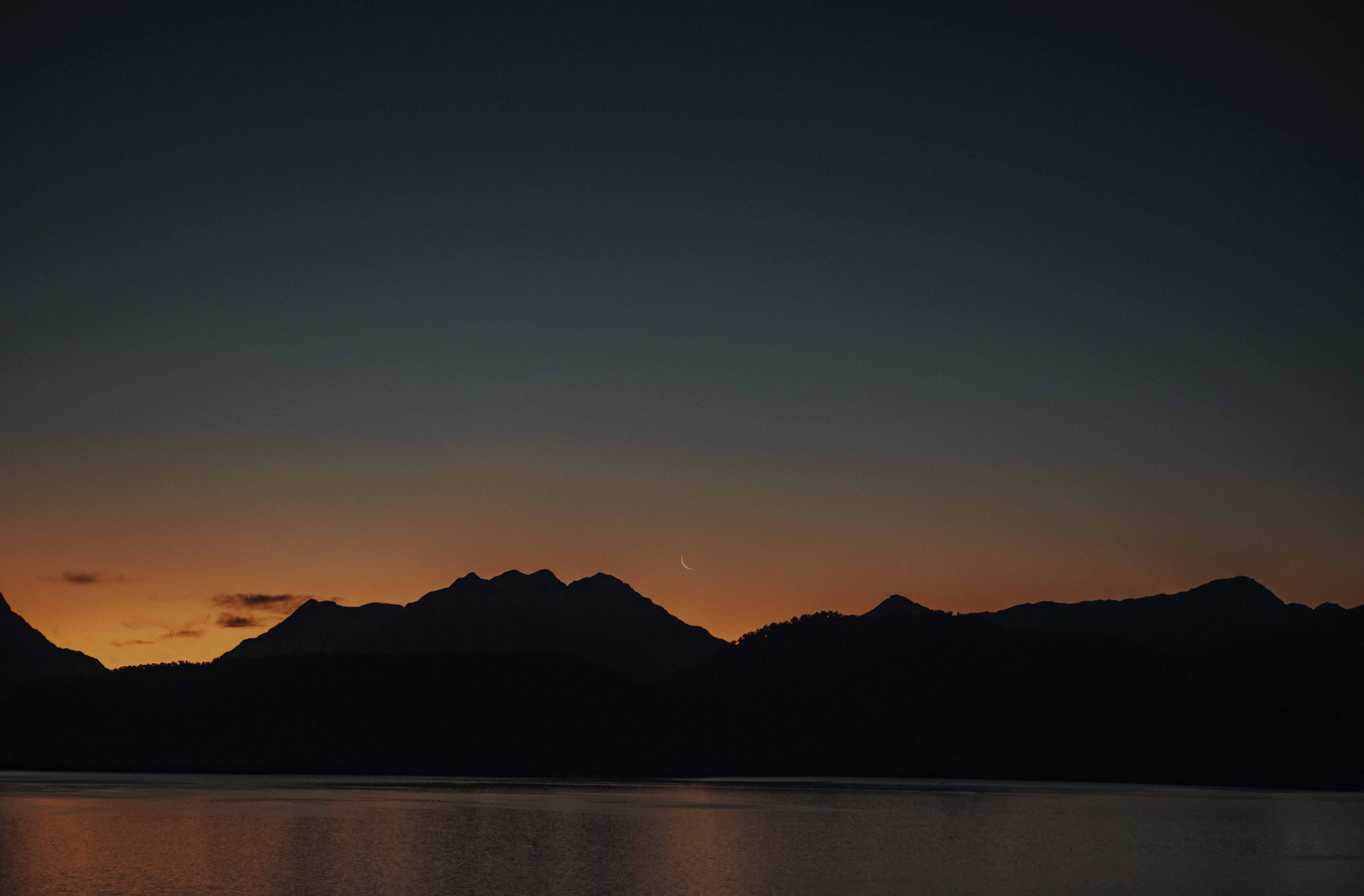 A nighttime landscape with mountains silhouetted against a sky transitioning from dark to light.