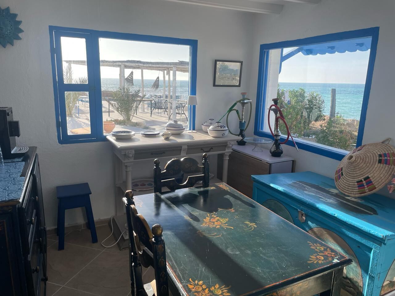 Une pièce comprenant une table à manger, des chaises, un petit tabouret, un banc bleu, un narguilé et une vue sur la mer grâce à de grandes fenêtres.