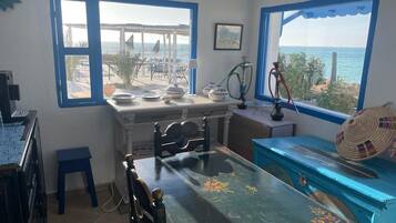 Une pièce comprenant une table à manger, des chaises, un petit tabouret, un banc bleu, un narguilé et une vue sur la mer grâce à de grandes fenêtres.