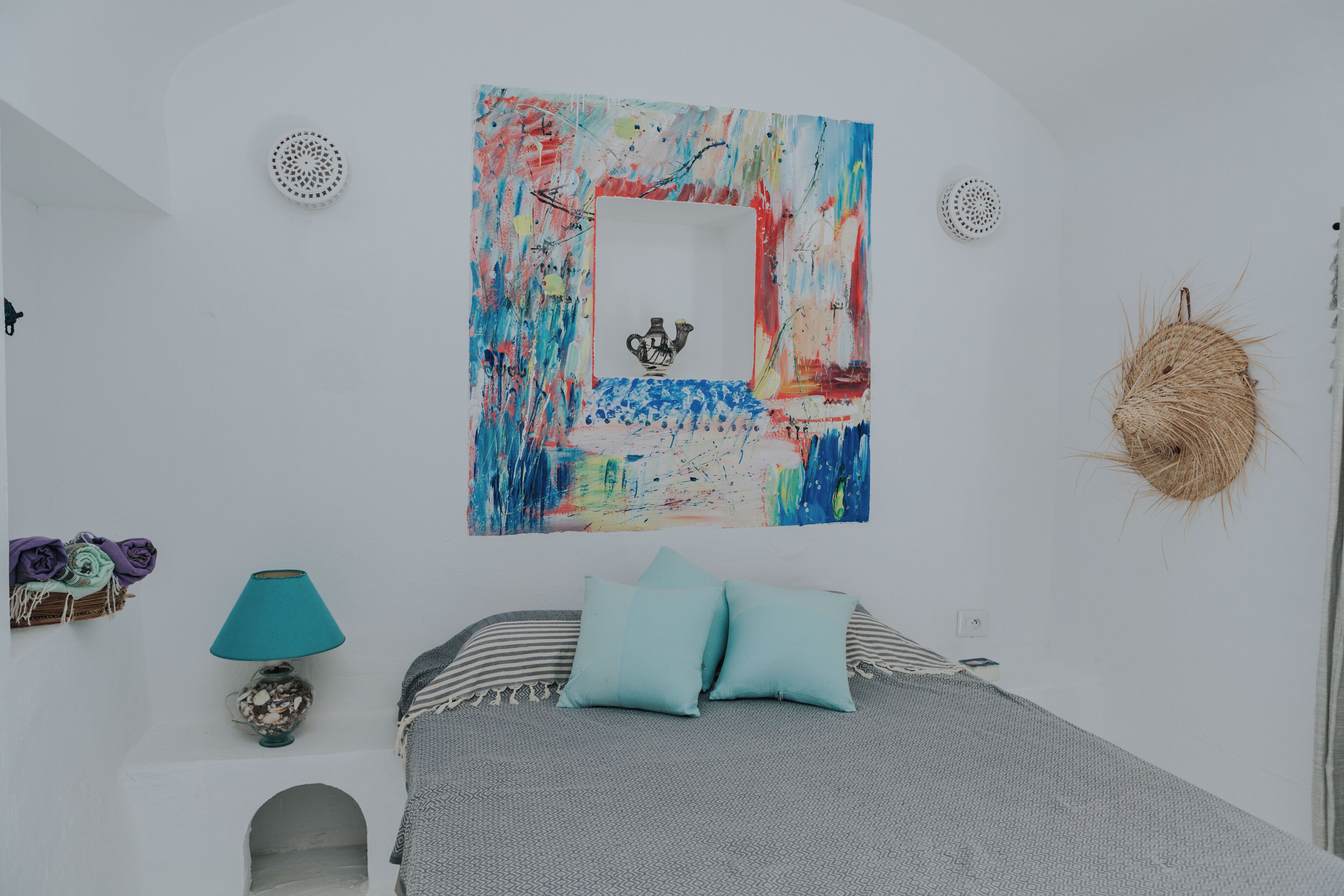 A bedroom with a bed, a wall-mounted artwork, a lamp, and a woven wall decoration.