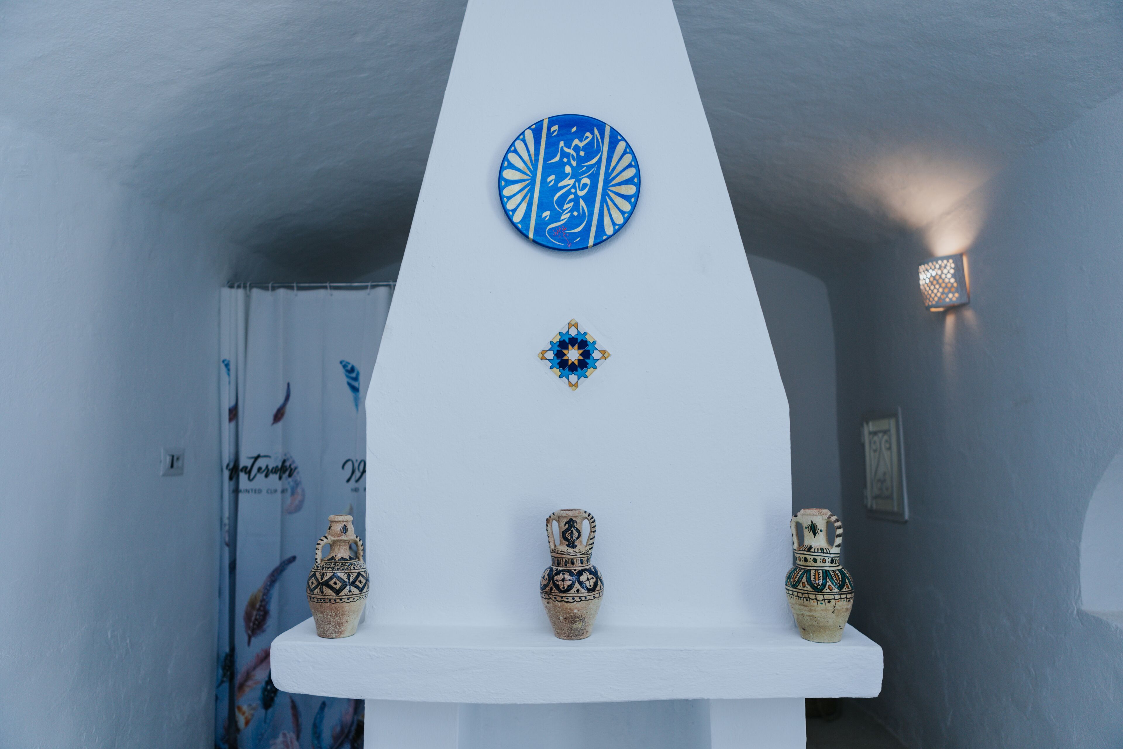 A white interior with a decorative blue plate, a small blue and gold ornament, and three patterned vases on a shelf.