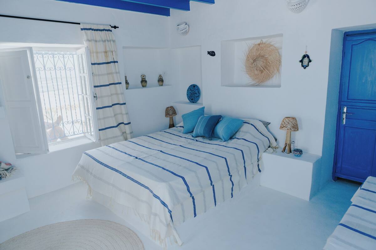 A bedroom with a bed, a window with curtains, a blue door, and wall decorations.