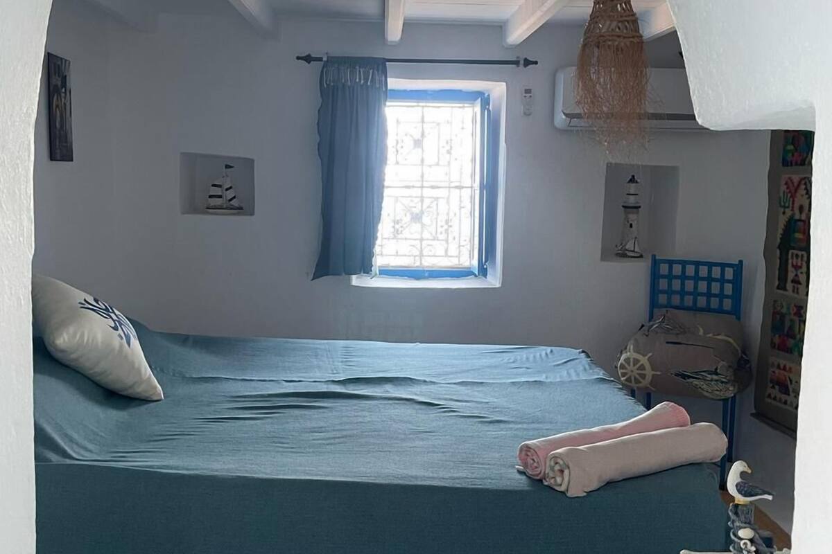 A bedroom with a single bed, a window with blue curtains, a hanging light fixture, and a small wall-mounted shelf.