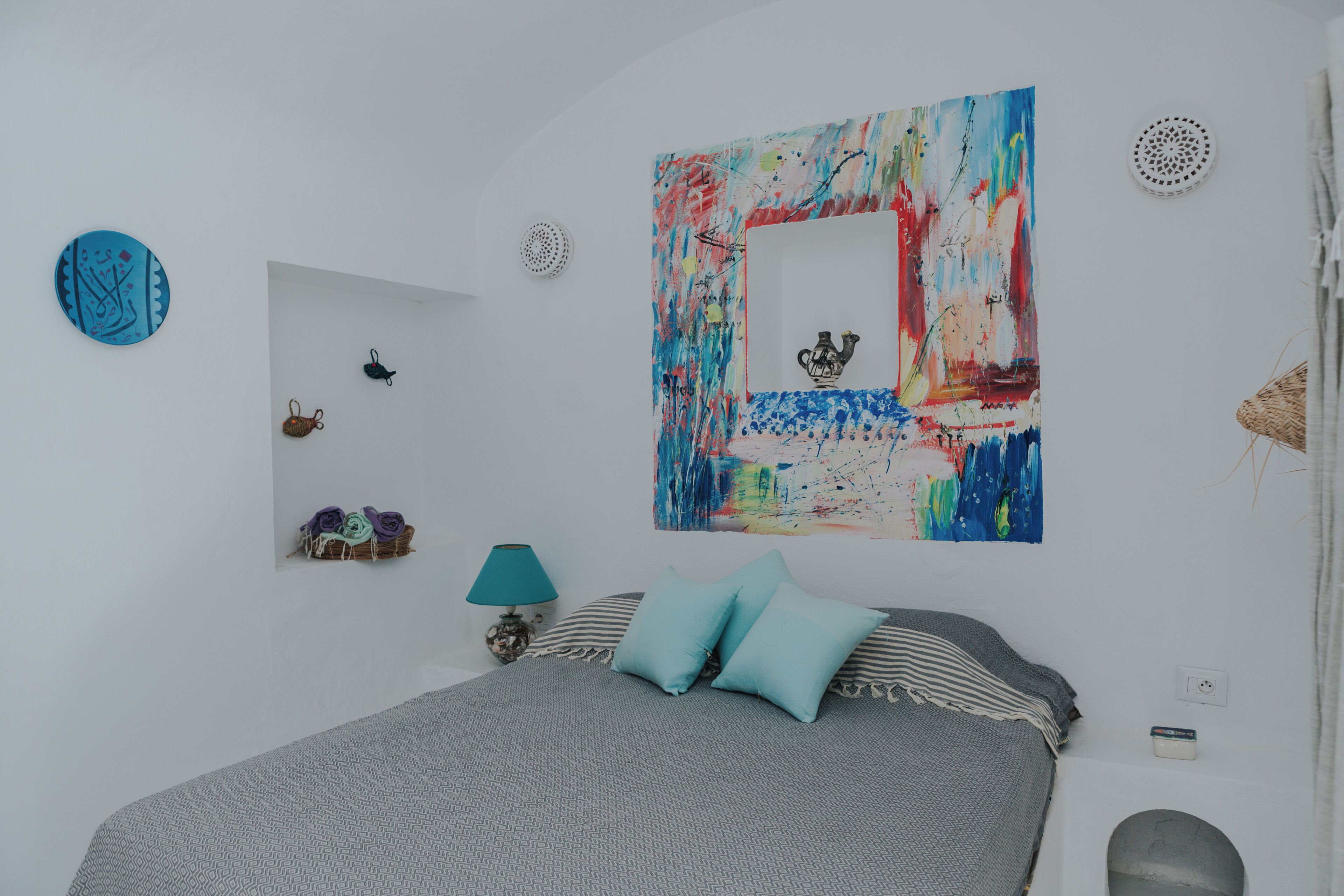 A bedroom with a bed, pillows, a lamp, a wall art piece, and a small shelf with decorative items.