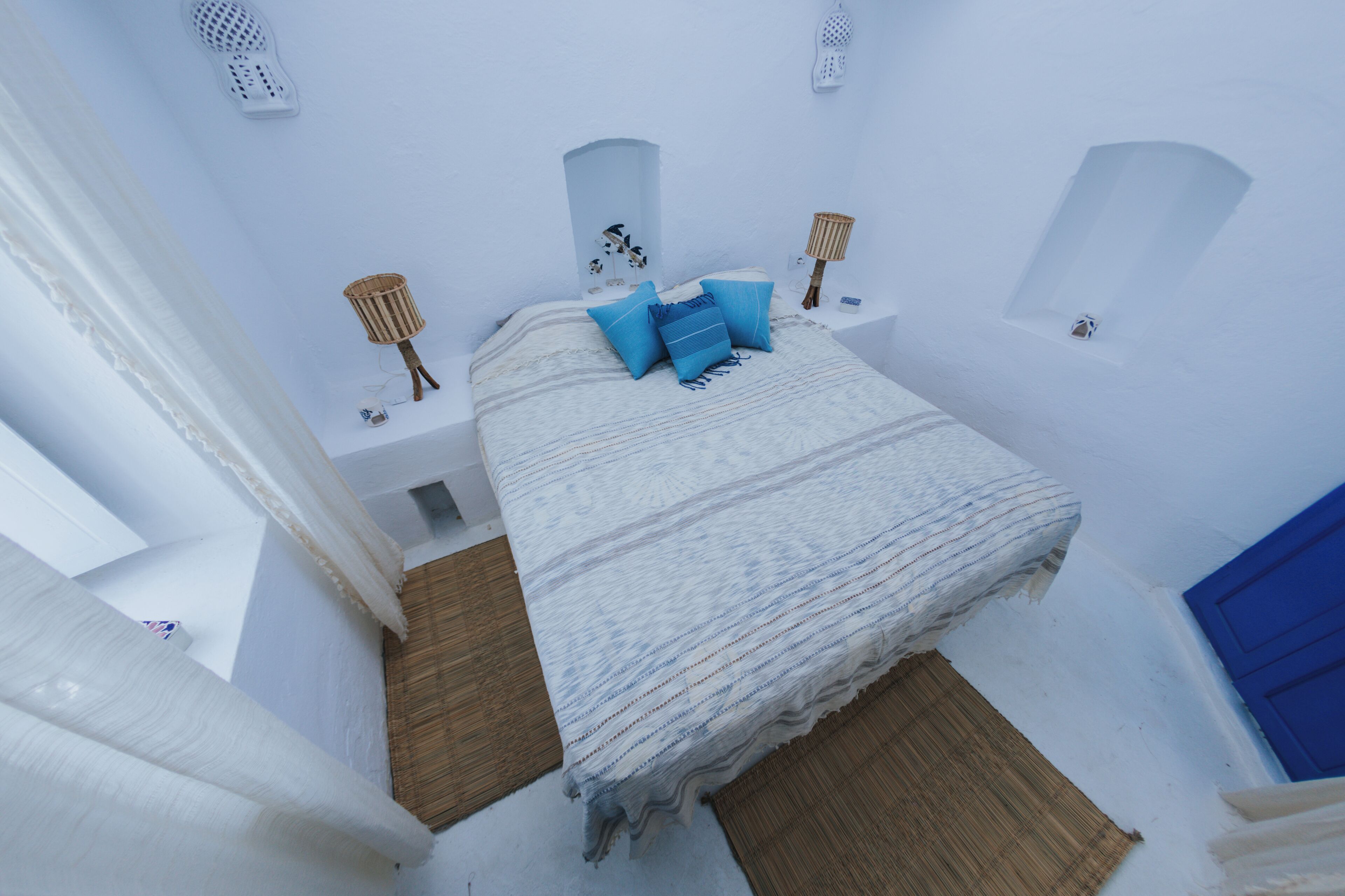 Een slaapkamer met witte wanden, een bed, kussens, een lamp en een blauwe deur.