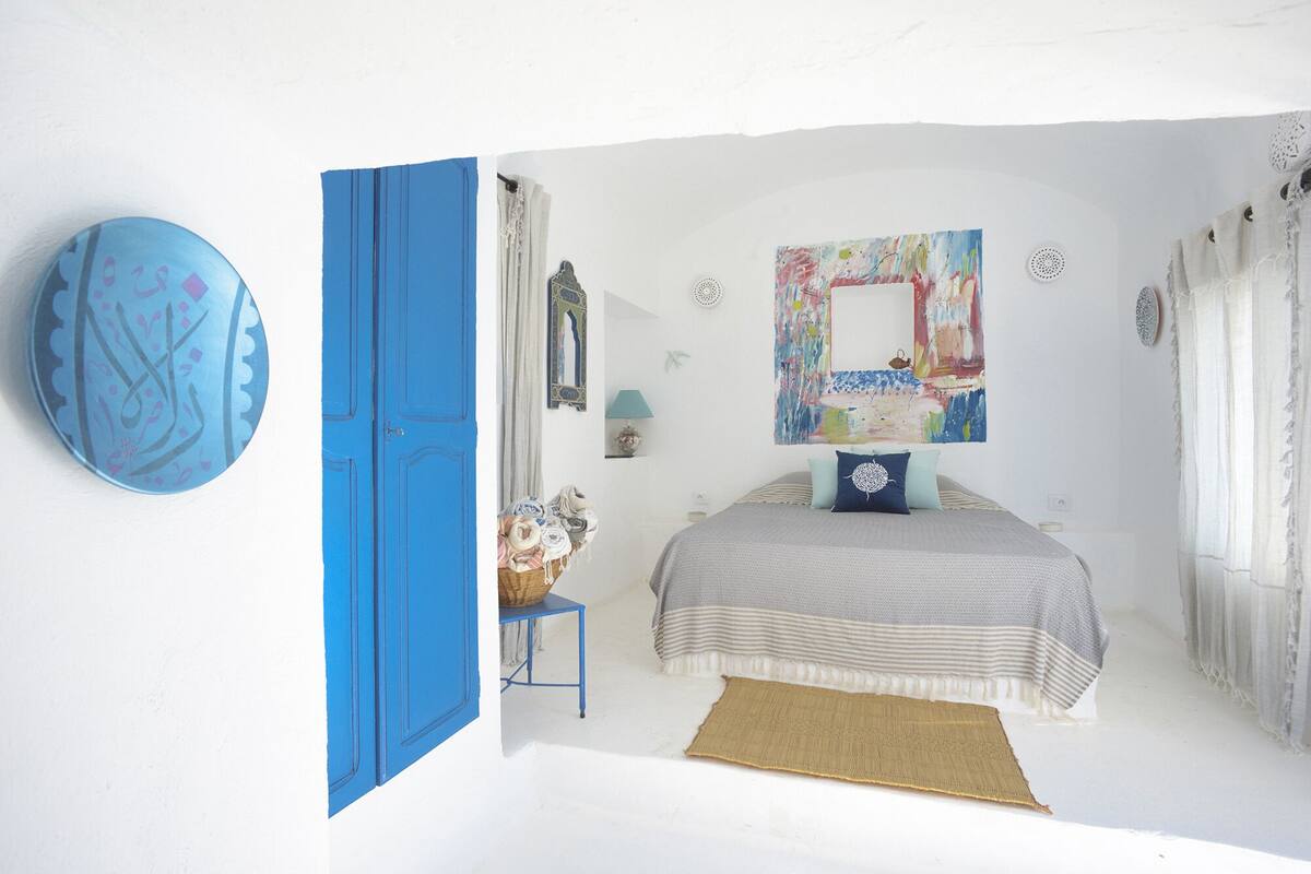 A bedroom with a bed, a blue door, a wall with a colorful painting, and a small table with a basket.