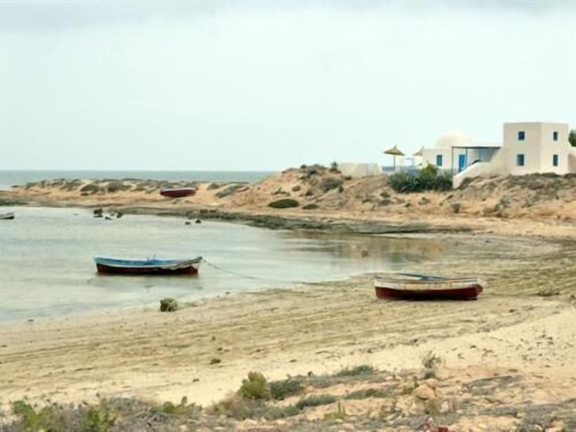 Escena costera con embarcaciones en la orilla, una playa arenosa y edificios de estilo arquitectónico sencillo.