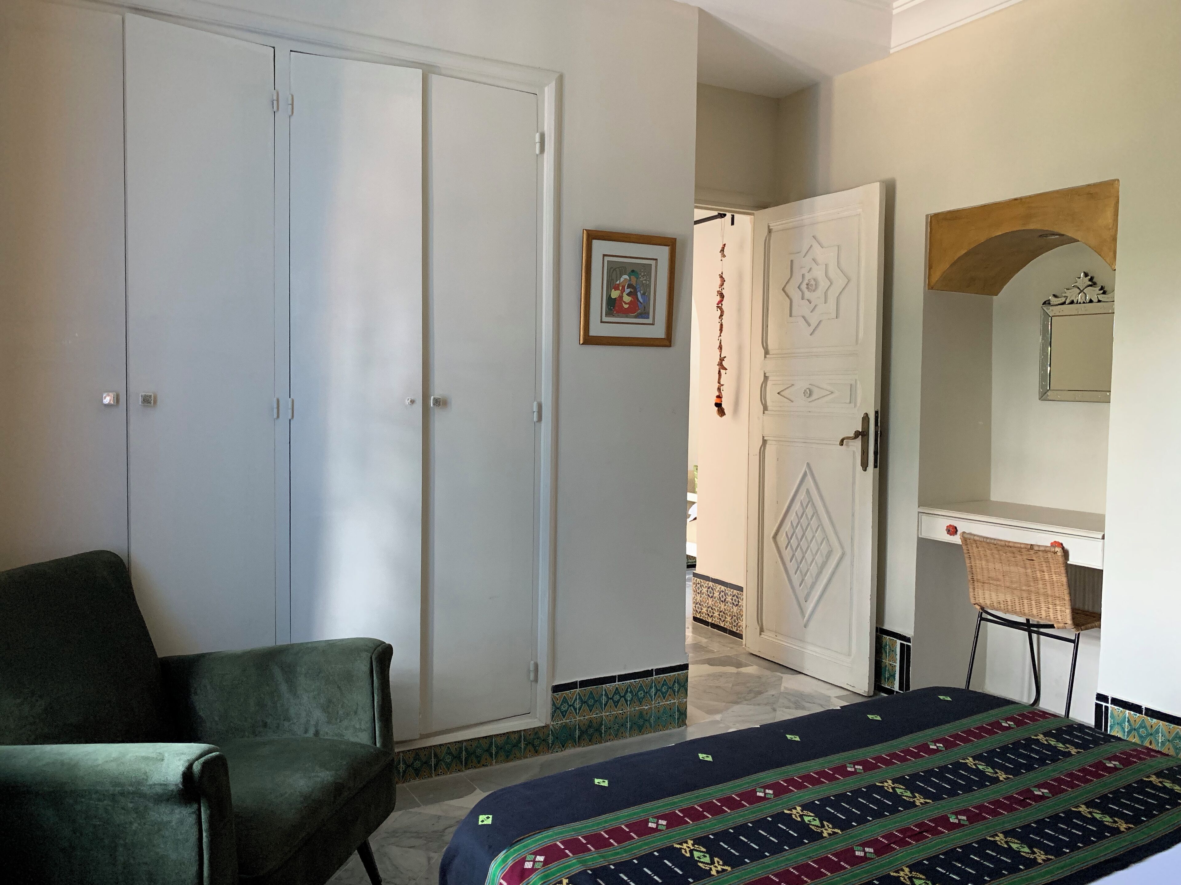 A room with a green armchair, a bed with a patterned bedspread, a white wardrobe, and a small desk with a chair.