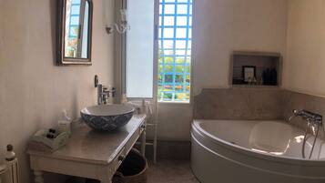 Un baño con una bañera grande, un lavamanos con un cuenco decorativo, un espejo y una ventana con vistas a follaje.