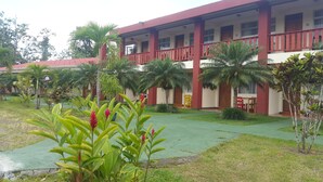 A building with a red exterior, multiple windows, and a balcony. There are palm trees and other greenery in the surrounding area. - Hotel Fuego Arenal (La Fortuna)
