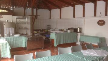 Un espace repas avec des tables recouvertes de nappes à carreaux verts, des chaises blanches et un coin cuisine avec des murs carrelés de blanc et des poutres en bois.