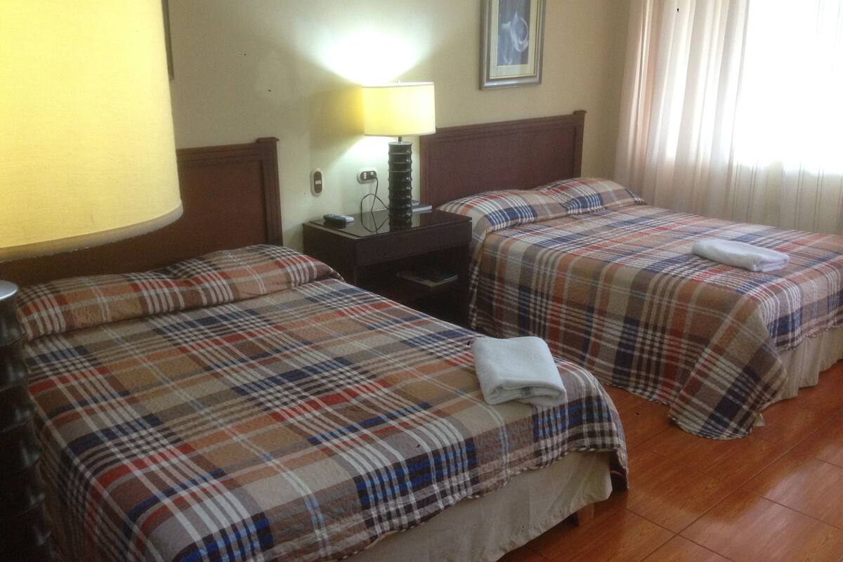 Habitación de hotel con dos camas, cada una con una colcha escocesa, una mesita de noche con una lámpara y un cuadro en la pared.