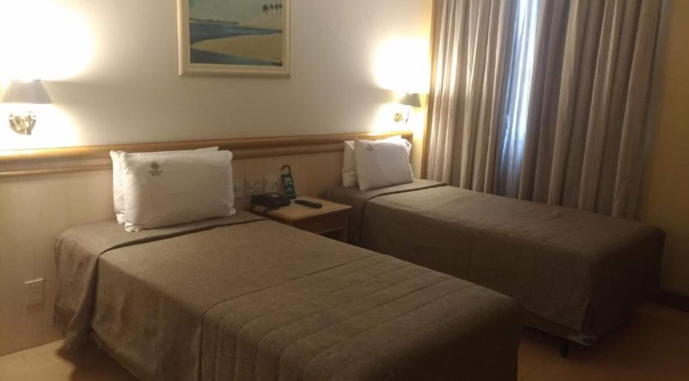 A hotel room with two single beds, a nightstand with a lamp, a painting on the wall, and a window with curtains.