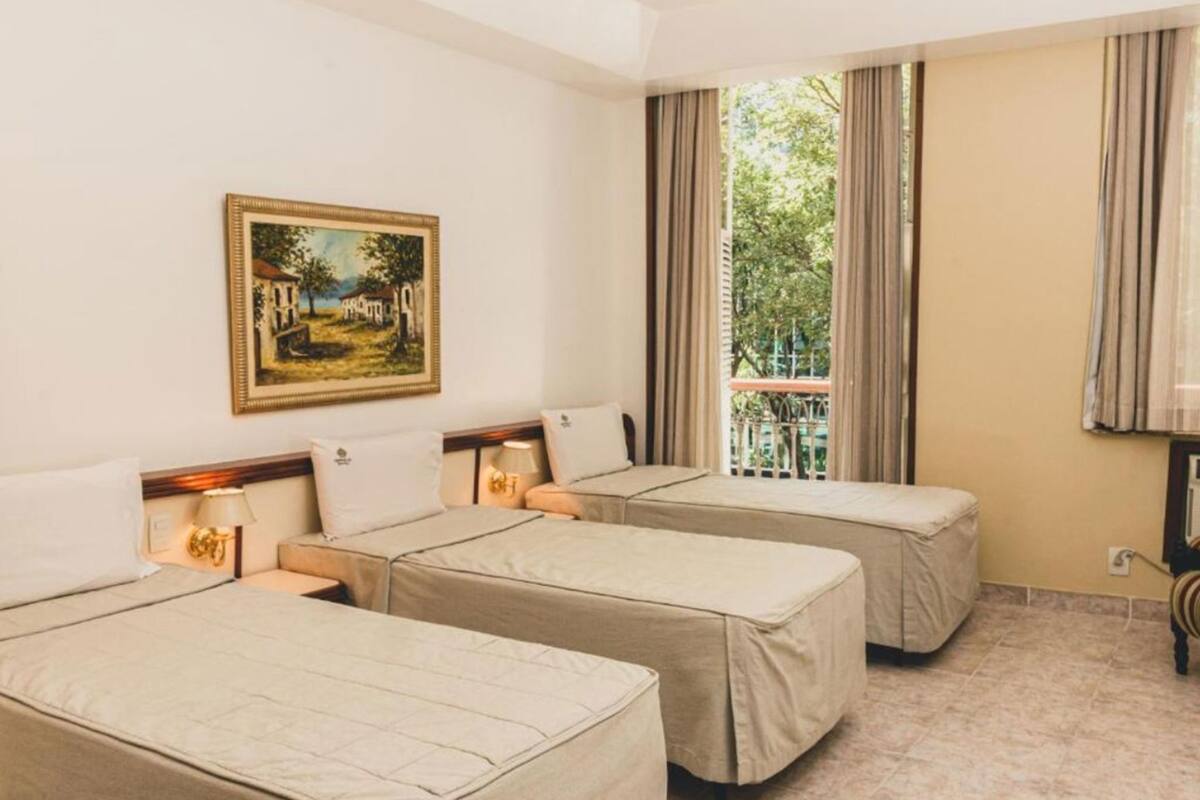 Ein Hotelzimmer mit zwei Betten, einem Bild an der Wand, einem Fenster mit Vorhängen und Blick auf Grünfläche.