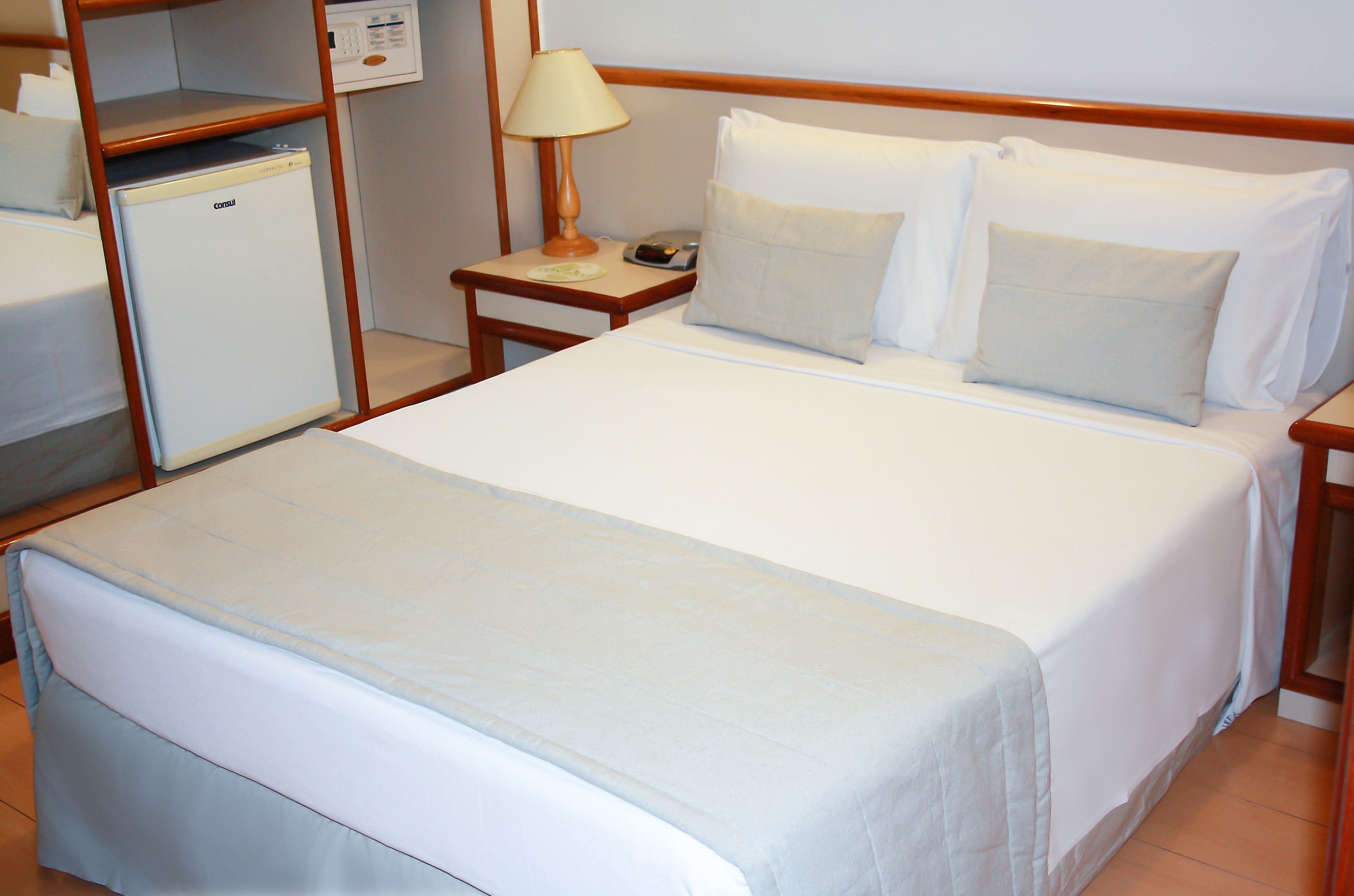 A hotel room with a single bed, a nightstand with a lamp, a small refrigerator, and a wall-mounted control panel.