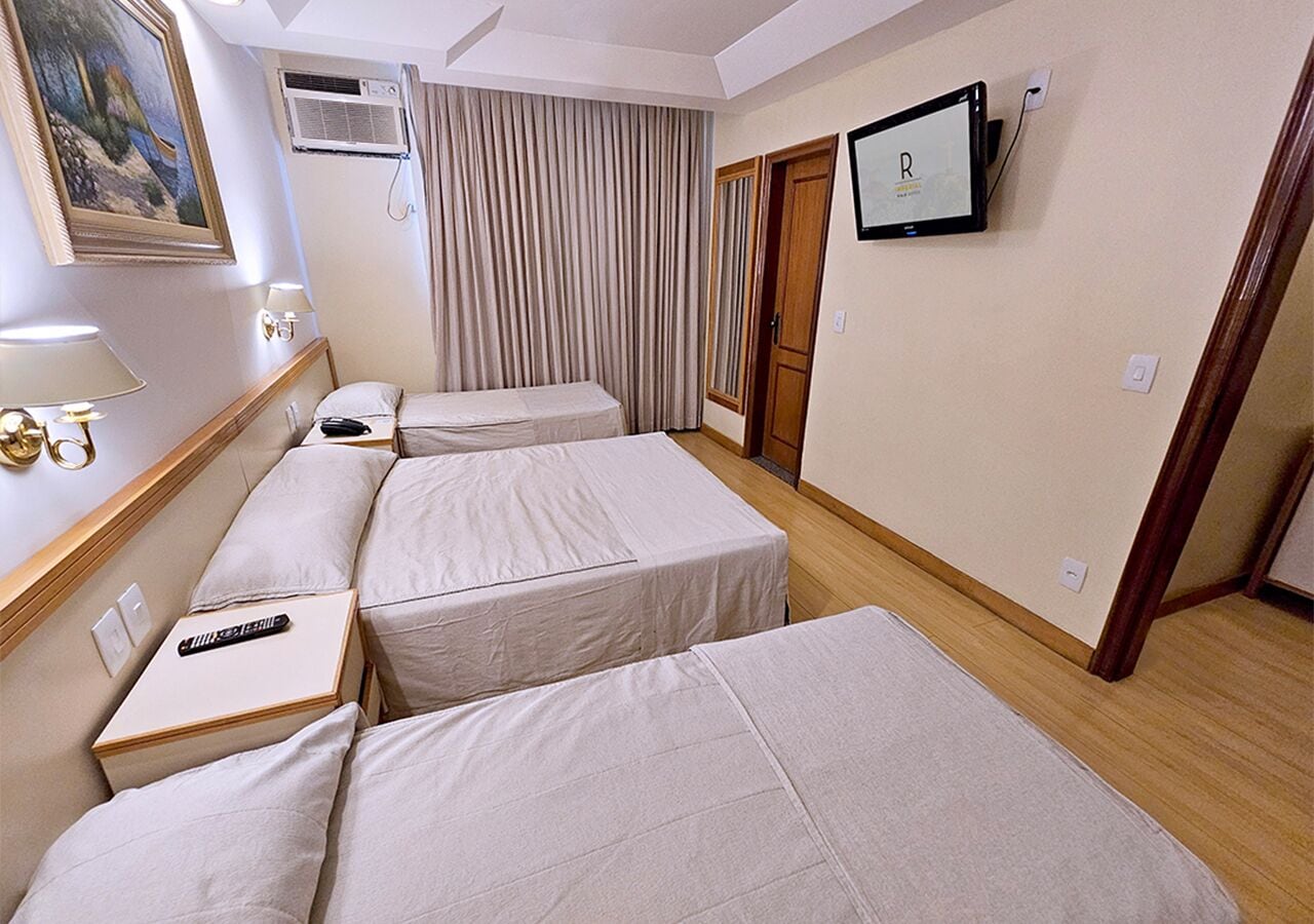 Habitación de hotel con tres camas individuales, un televisor montado en la pared, un cuadro en la pared y una ventana con cortinas.
