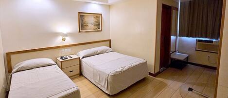 A hotel room with two beds, a bedside table with a lamp, a painting on the wall, and a small seating area with a chair and table.