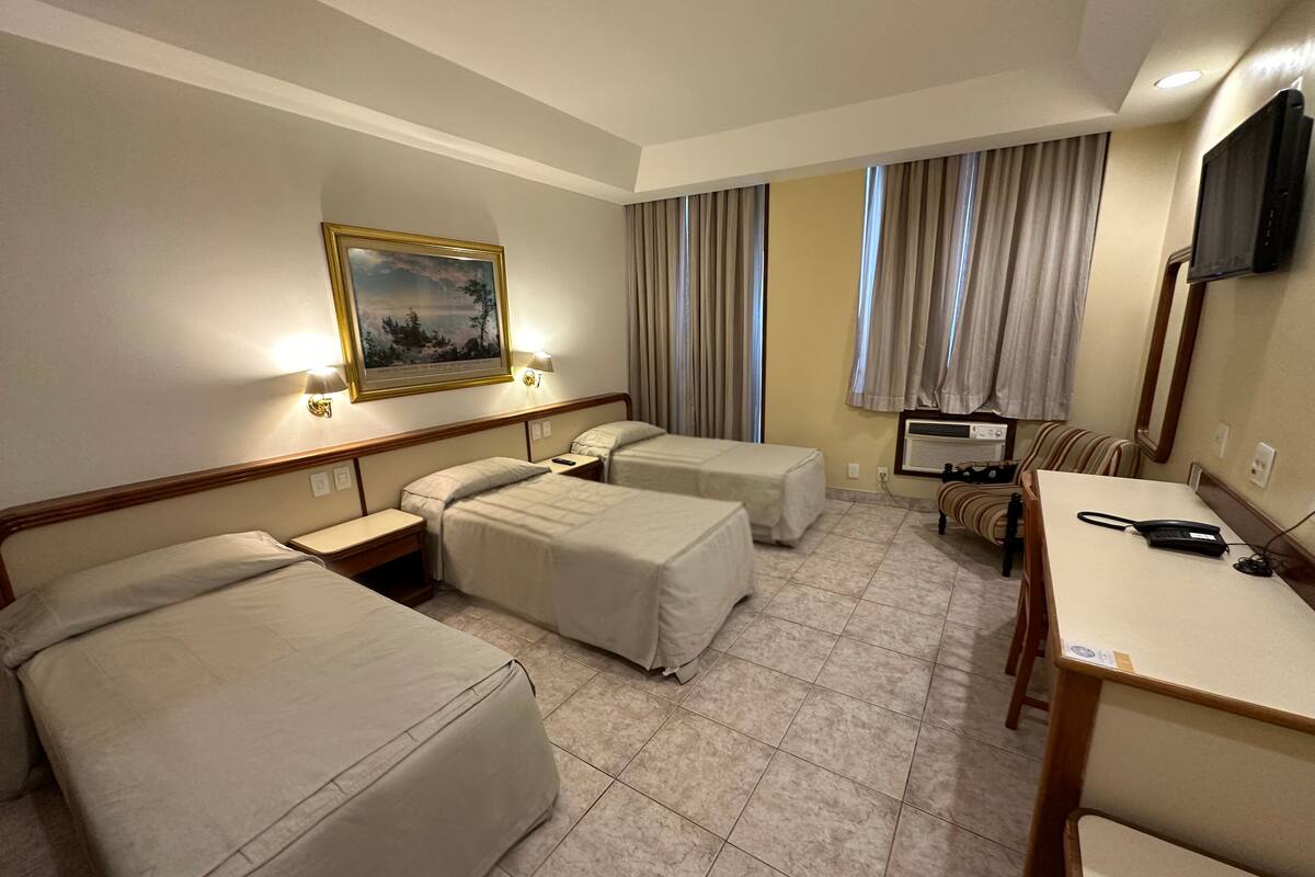 Ein Hotelzimmer mit zwei Einzelbetten, einem kleinen Tisch mit Telefon, einem Sessel, einem an der Wand befestigten Fernseher und einem Gemälde über den Betten.