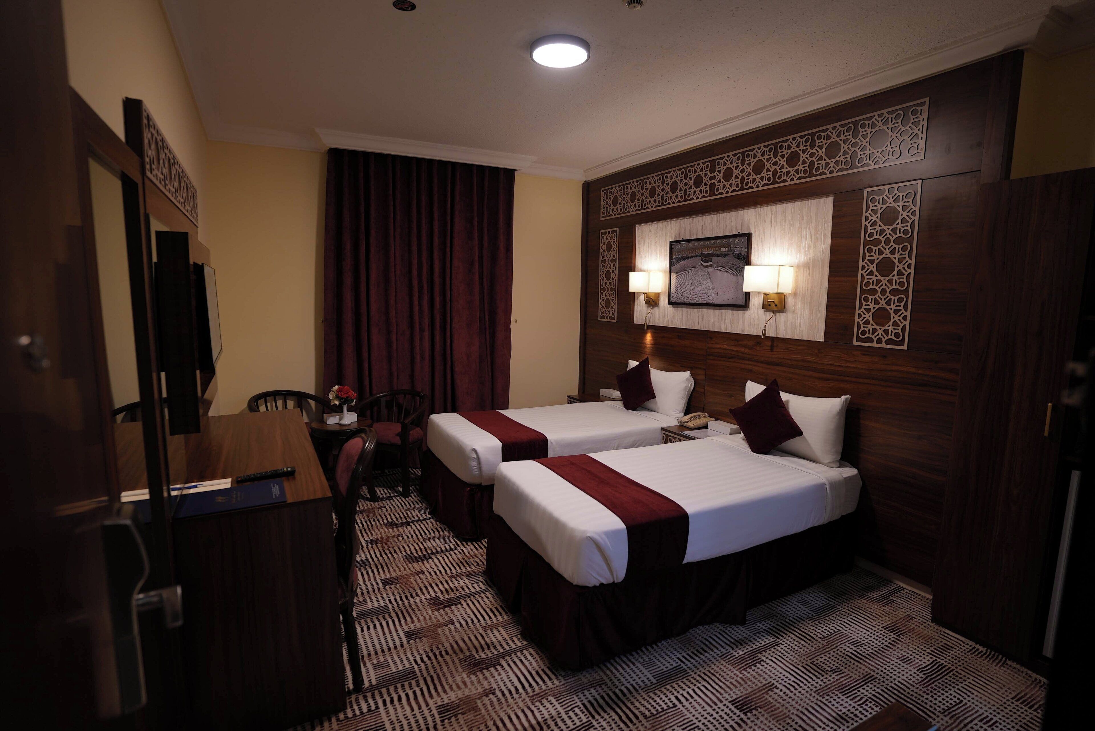 Quarto de hotel com duas camas, uma escrivaninha, uma cadeira, uma televisão e um tapete estampado.