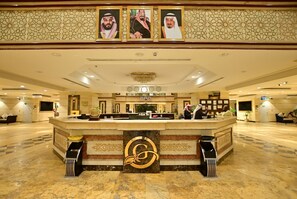 Een hotel receptie met een marmeren vloer, een receptiebalie en drie ingelijste portretten erboven.