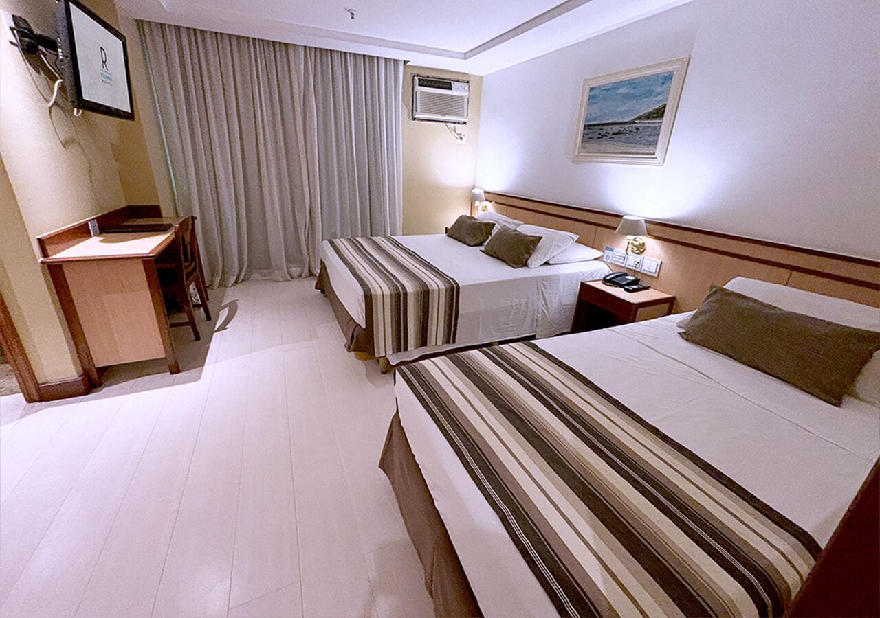 A hotel room with two single beds, a desk, a chair, a nightstand with a lamp, and an air conditioning unit.