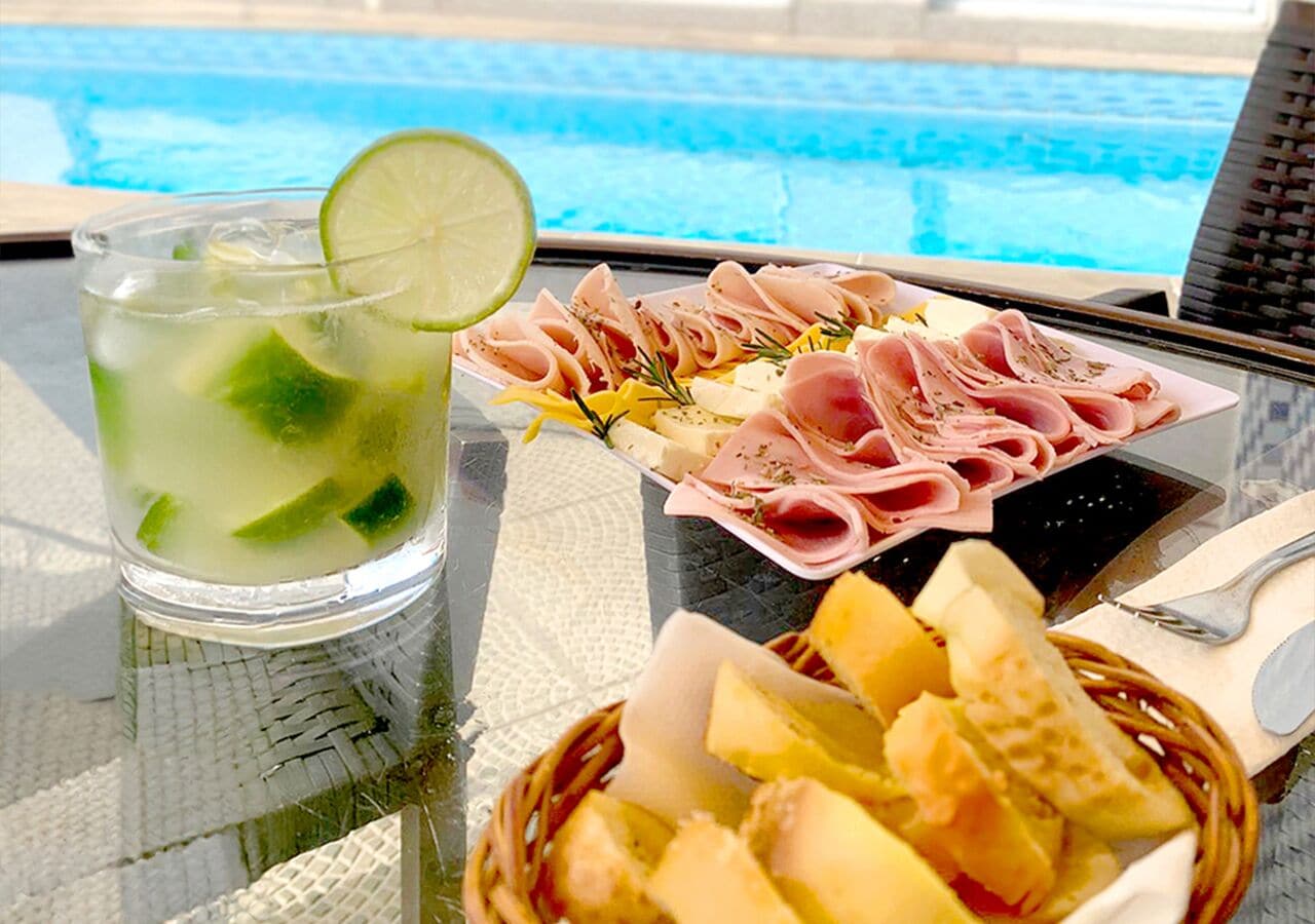 Uma mesa com um copo de limonada, um prato de charcutaria e uma cesta de fatias de queijo.