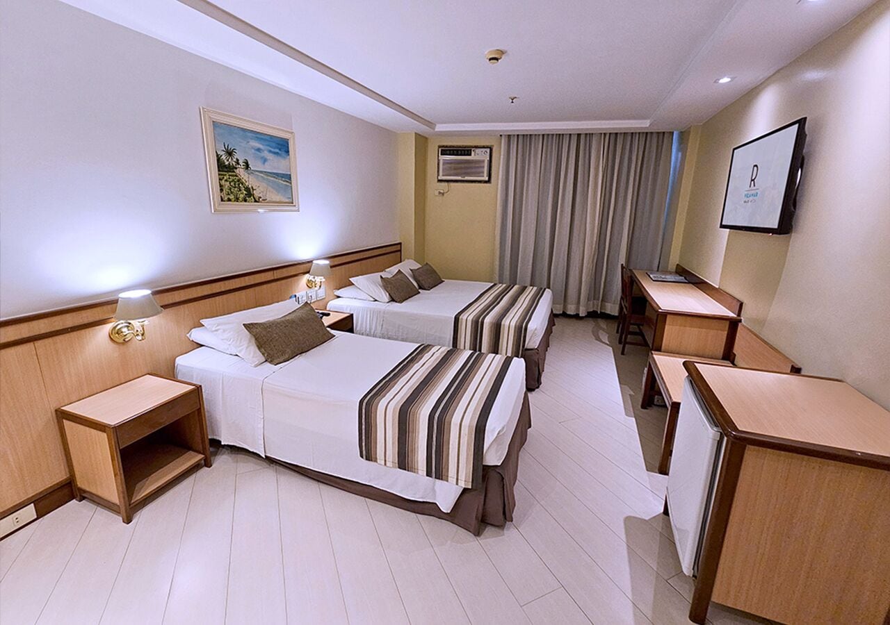Quarto de hotel com duas camas, uma escrivaninha, uma cadeira, uma mesa de cabeceira e uma televisão fixada na parede.