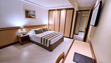 Quarto de hotel com cama, mesa de cabeceira, luminária e escrivaninha com cadeira.
