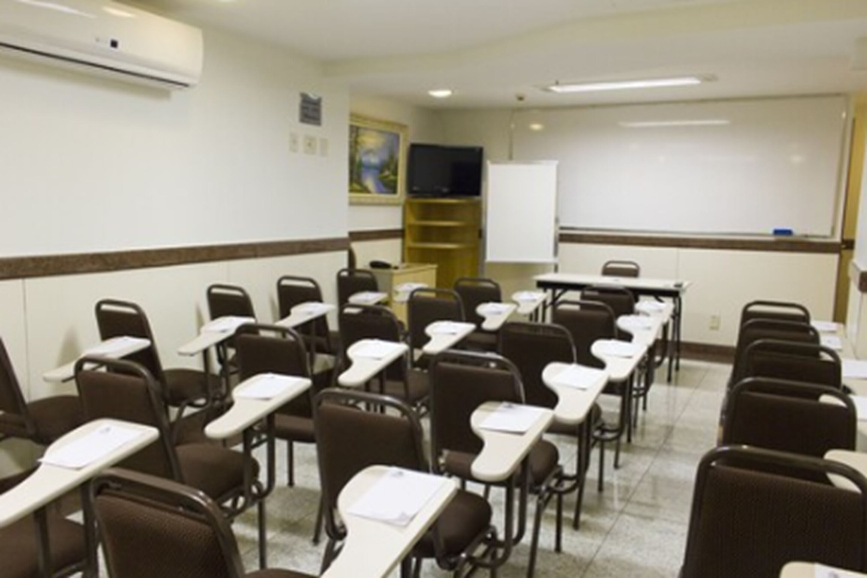 Uma sala de aula com fileiras de mesas e cadeiras, um quadro branco e uma televisão em uma prateleira.