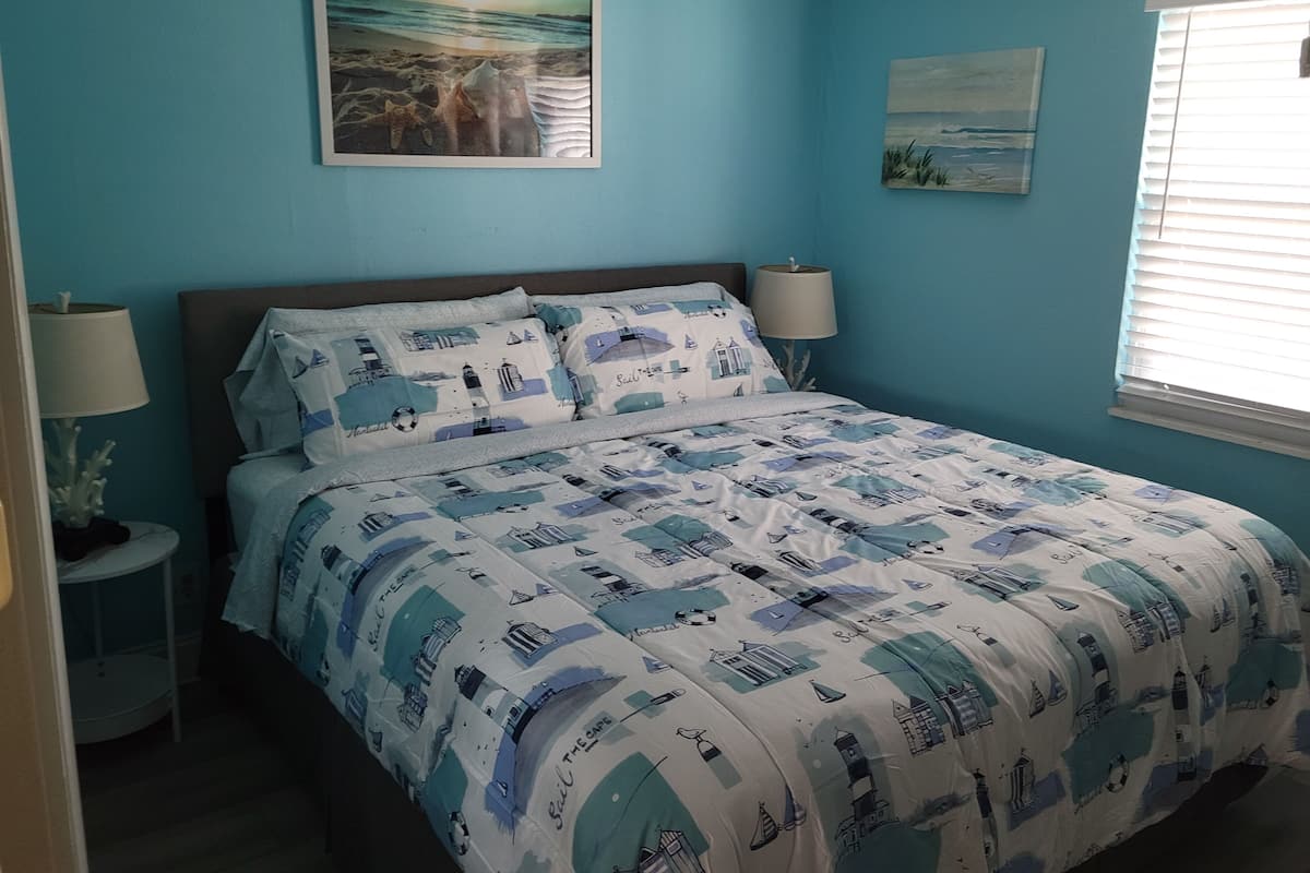 A bedroom with a bed, two bedside tables, a nightstand, a window with blinds, and two wall-mounted lamps.
