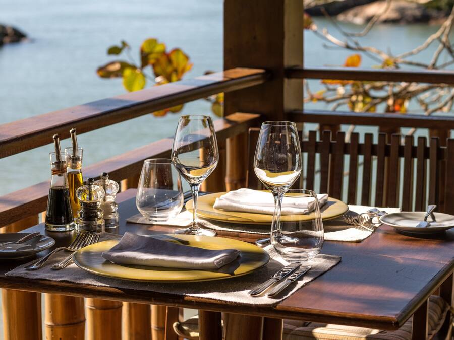 Mesa lista para cenar con copas de vino, platos y cubiertos, con vista a un cuerpo de agua.