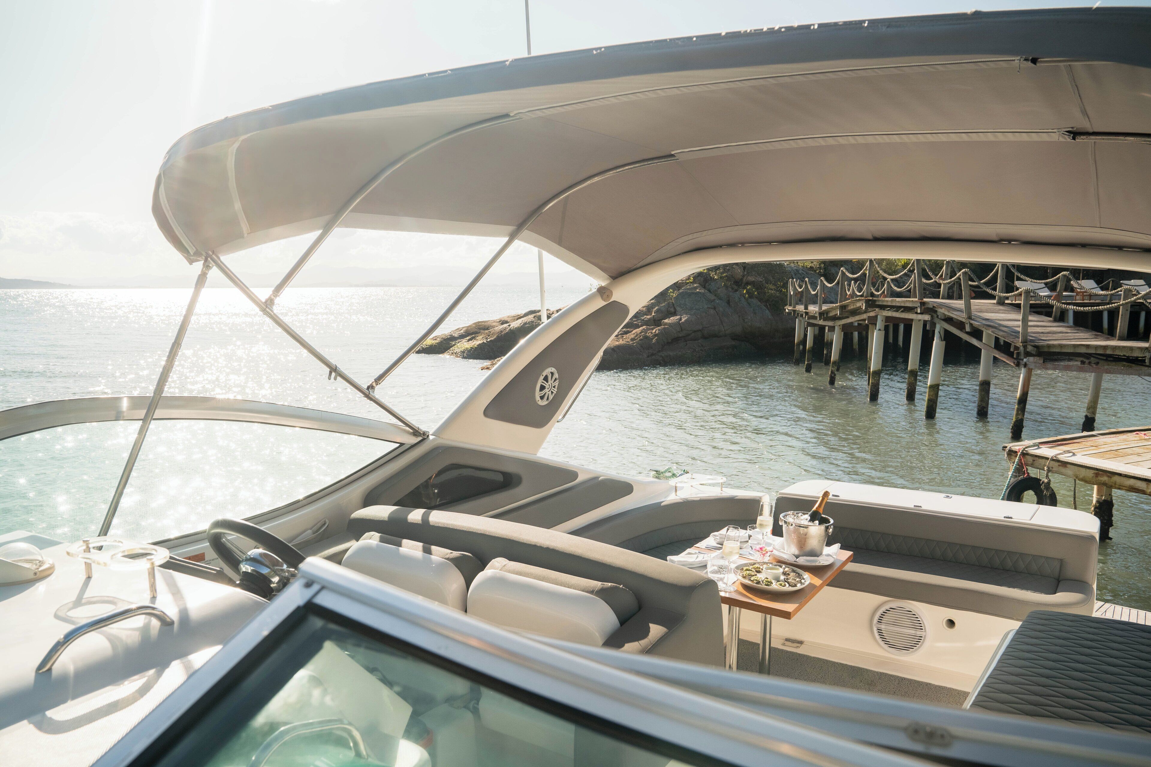 A boat's interior with a dining table, cushioned seating, and a view of a pier.