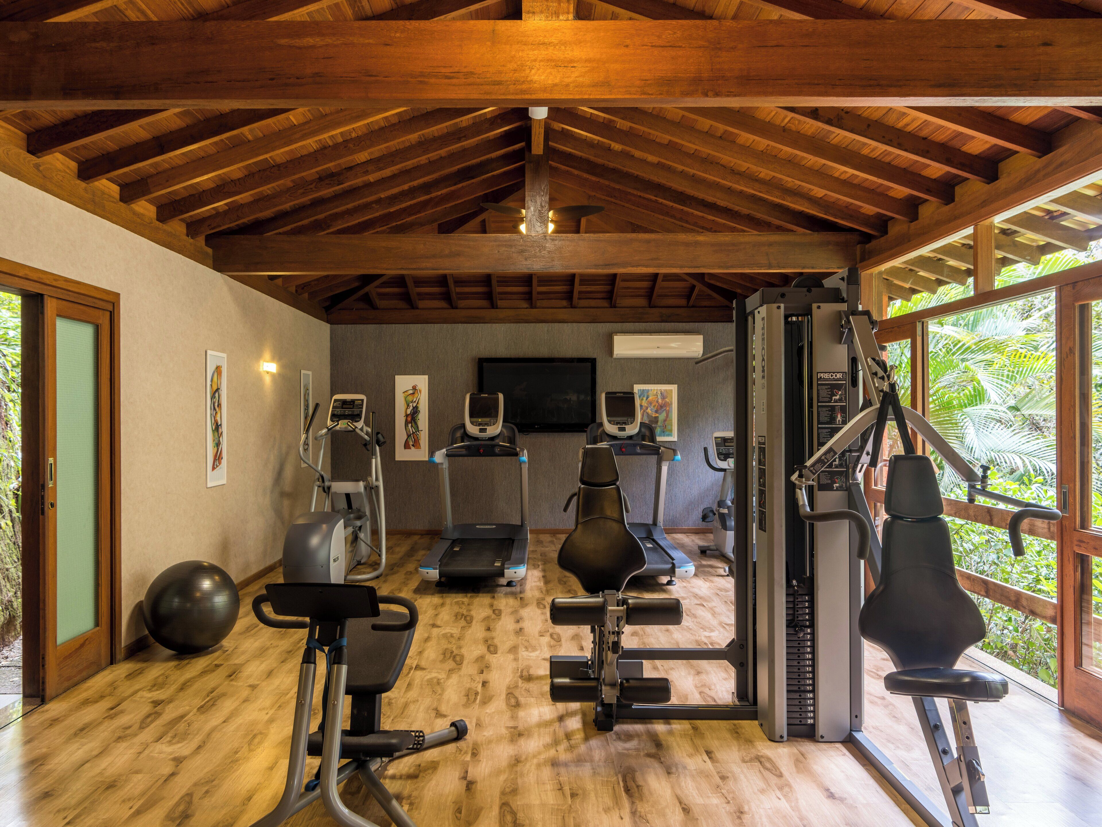 Ein Fitnessraum mit verschiedenen Trainingsgeräten, einem Fernseher und Blick auf Grünflächen durch große Fenster.