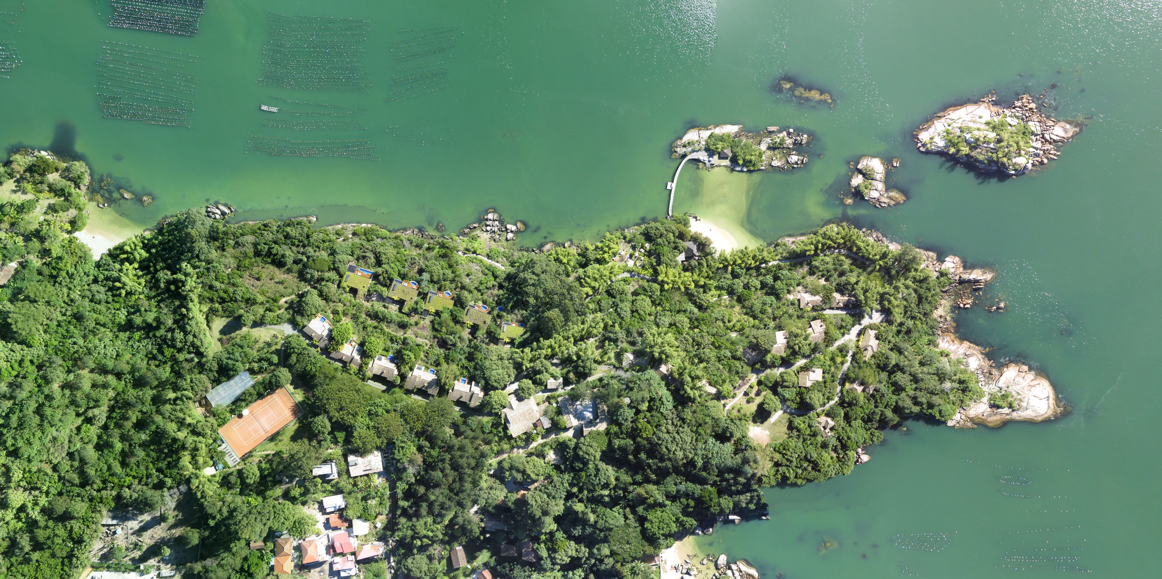 An aerial view of a coastal area with dense greenery, scattered buildings, and a clear water body.