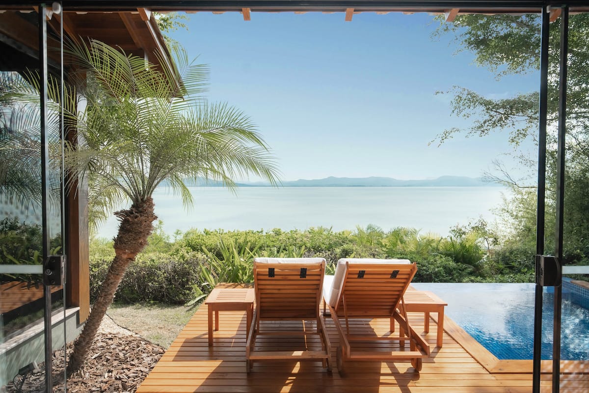 A wooden deck with two lounge chairs facing a pool and a view of a lake.