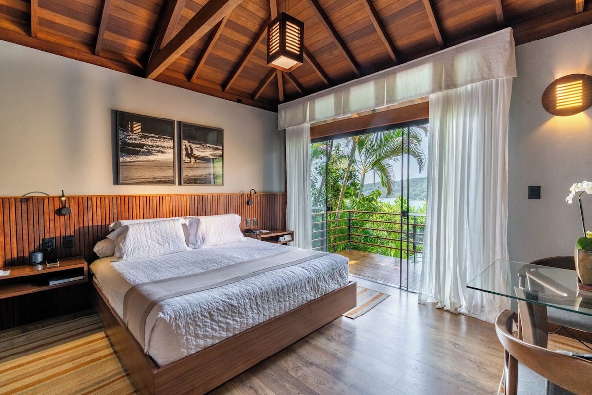 A bedroom with a large bed, wooden ceiling beams, a nightstand, a glass table, and a view of palm trees through a sliding glass door.