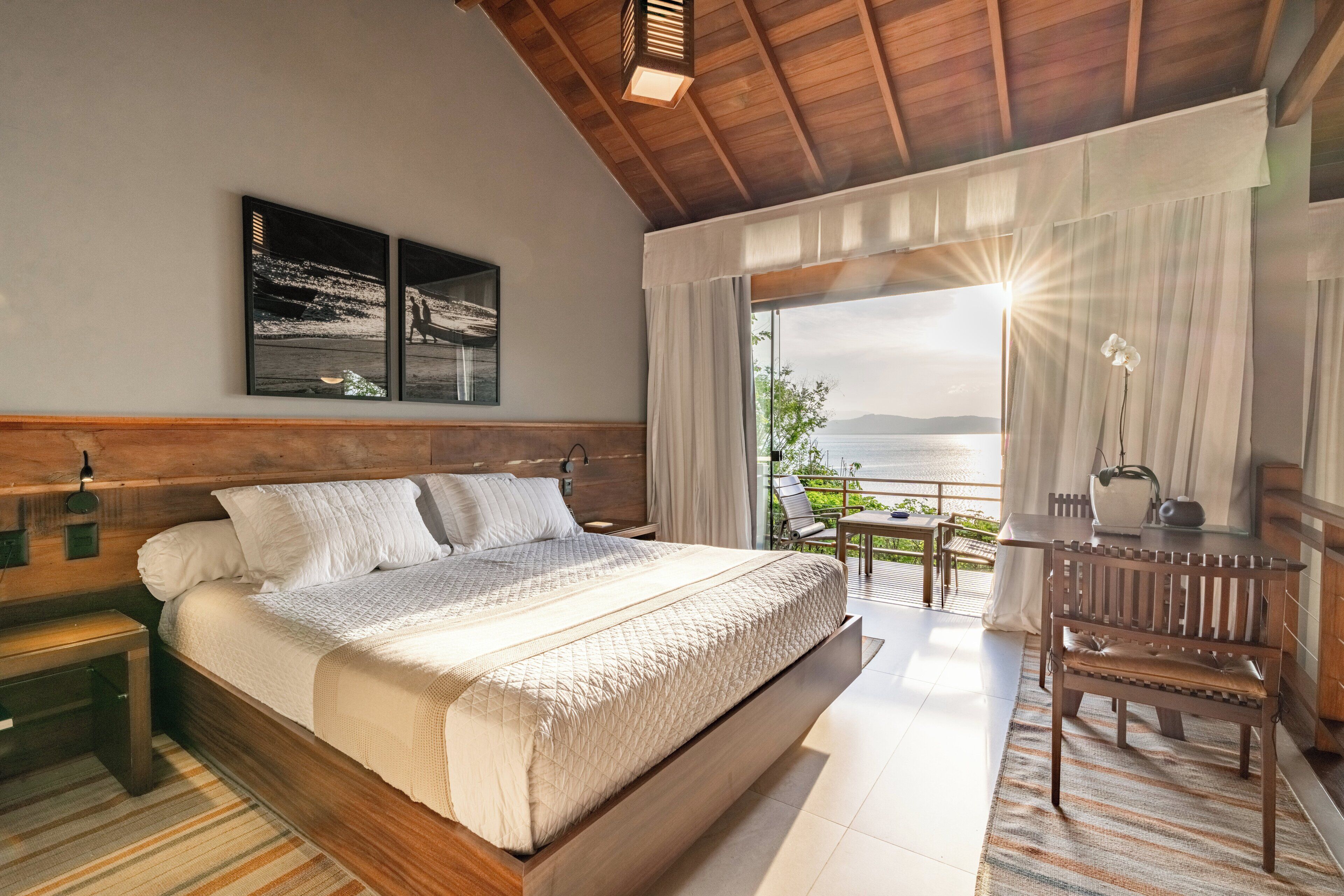 Une chambre moderne avec un grand lit, une tête de lit en bois, une table de chevet, un banc, un bureau avec une chaise et une vue sur la mer grâce à de grandes fenêtres.