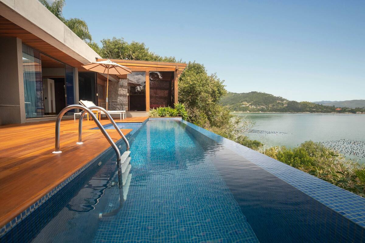 A modern pool with a wooden deck and glass doors, overlooking a lake.