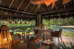 A room with a thatched roof, a dining table set for two, and a view of a beach and water.